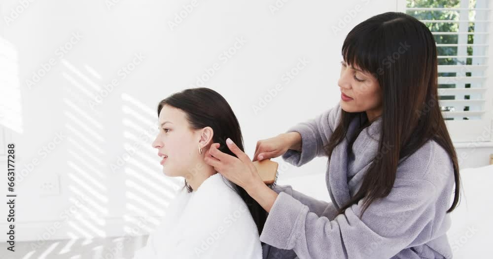 Happy biracial mother in bathrobe brushing hair of smiling adult daughter in sunny room, slow motion Happy biracial mother in bathrobe brushing hair of smiling adult daughter in sunny room, slow motion