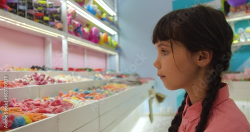 Portrait of little European girl with brown hair braided and wearing a pink shirt choosing and buying candy in a store.