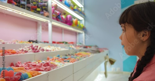 Portrait of little European girl with brown hair braided and wearing a pink shirt choosing and buying candy in a store.