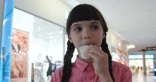 Portrait of little European girl with brown hair braided and wearing a pink shirt eating candy in a store.