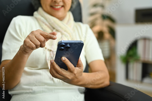 Pleasant senior woman using smartphone feeling satisfied sending messages while sitting on armchair at home