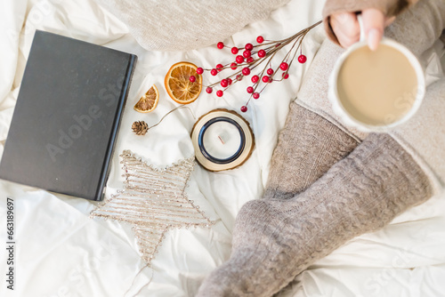 Cozy winter scene. Christmas vibes. Woolen socks. Woman having coffee in bed with book and candle.