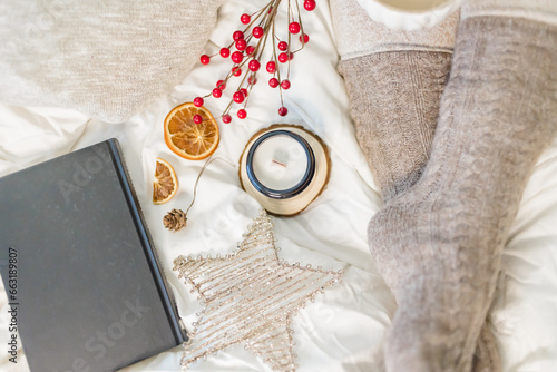 Cozy winter scene. Christmas vibes. Woolen socks. Woman having coffee in bed with book and candle.
