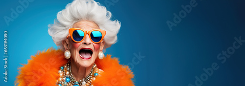 Happy senior woman grandmother in colorful neon outfit, funny sunglasses and extravagant style, laughing and smiling, trendy grandma posing in studio. With copy space.