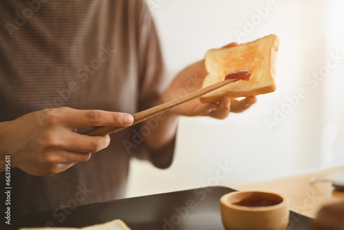 Young man preparing a healthy breakfast spreading strawberry jam on slice of bread