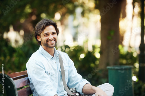 Travel, relax and portrait of a man on a park bench for a break, morning commute or summer. Smile, nature and a young person or tourist in a public garden for tourism, sightseeing or a vacation