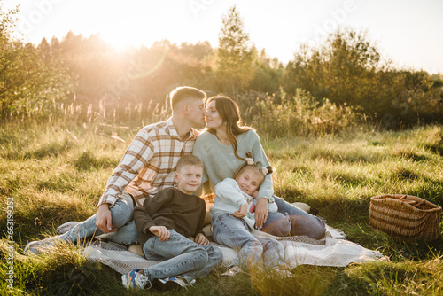 Mother, father, daughter, son sitting on blanket, hugging in grass in field at sunset. Family holiday outdoors. Children embrace parents. Happy young family walking spending time together in nature.