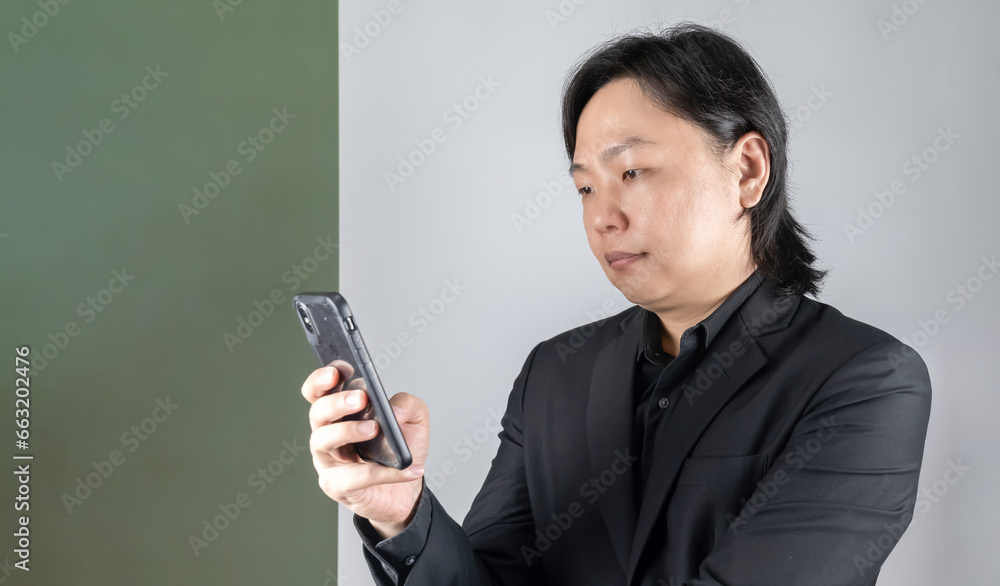 The portrait of a young Asian man dressed in a sleek black suit, engrossed in his mobile device