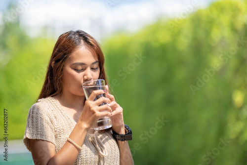 Asian Woman drinks water from tall glass of water at outdoor tree bokeh green background. photo