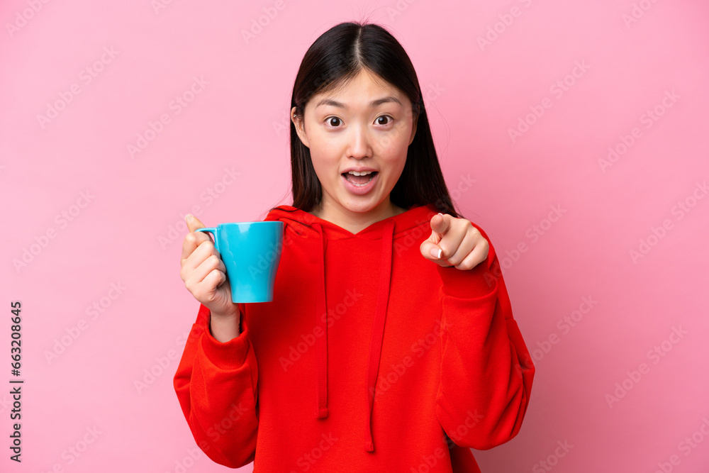Young Chinese woman holding cup of coffee isolated on pink background surprised and pointing front Young Chinese woman holding cup of coffee isolated on pink background surprised and pointing front