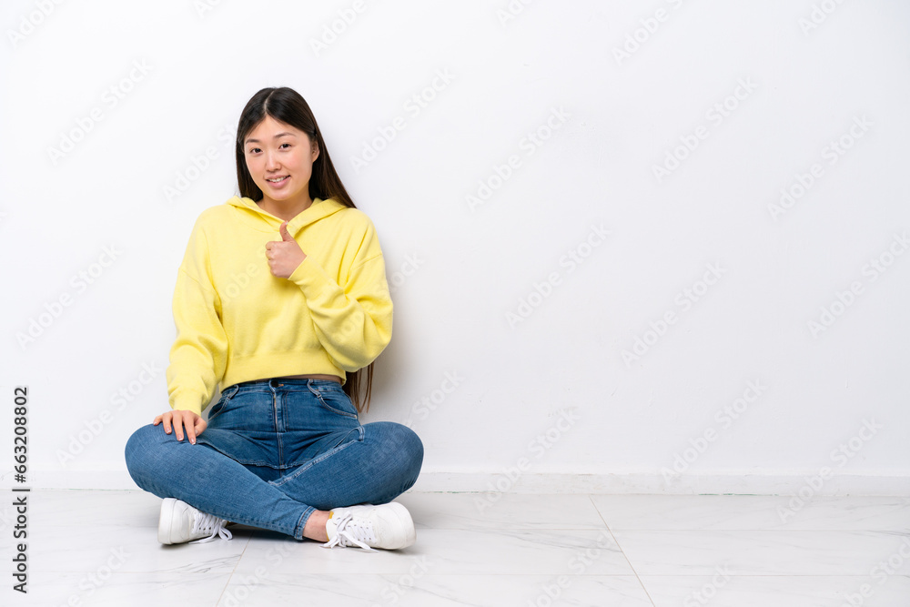 Young Chinese woman sitting on the floor isolated on white wall giving a thumbs up gesture Young Chinese woman sitting on the floor isolated on white wall giving a thumbs up gesture