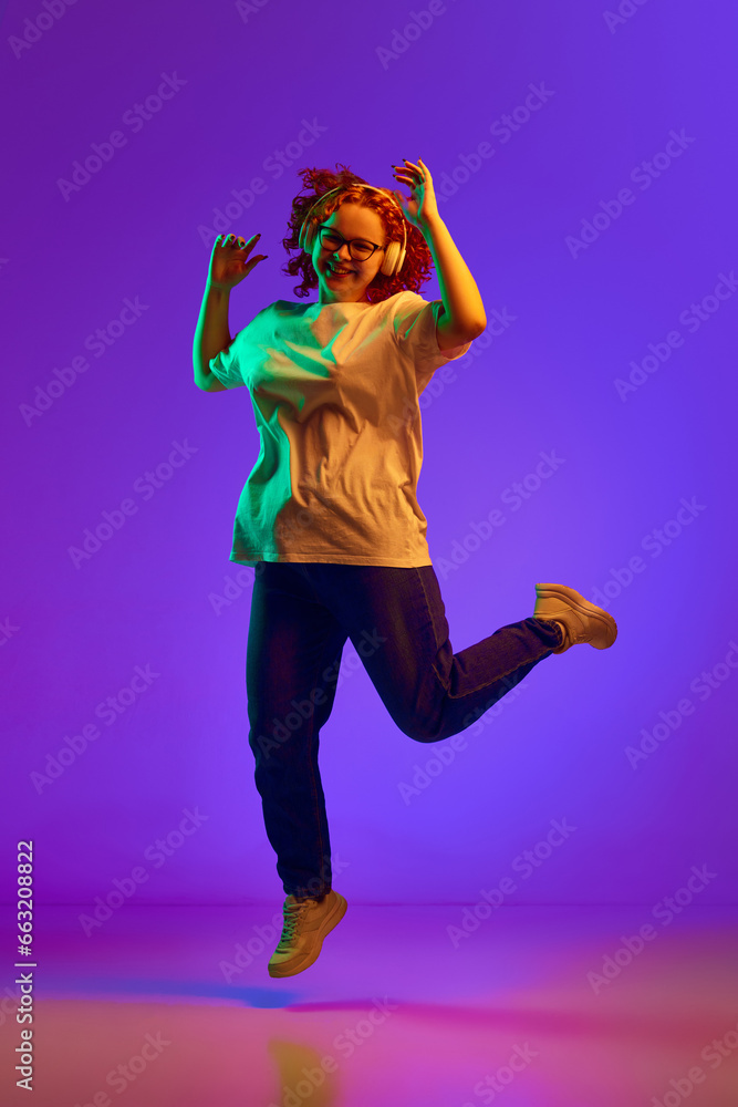 Happiness. Teen girl with curly hair listening to music in headphones and dancing against purple background in neon light. Concept of human emotions, lifestyle, youth, facial expression. Ad