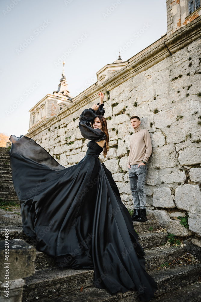 Luxury couple near old Pidhirtsi Castle, Lviv, Ukraine. Woman in black long dress with man stand on stairs near ancient palace at sunset. Female and male wall in street. Bride with groom outdoors