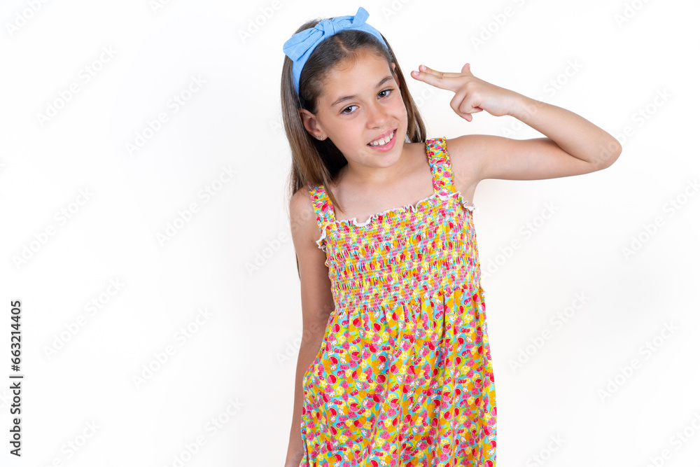 Beautiful kid girl wearing floral dress foolishness around shoots in temple with fingers makes suicide gesture. Funny model makes finger gun pistol Beautiful kid girl wearing floral dress foolishness around shoots in temple with fingers makes suicide gesture. Funny model makes finger gun pistol