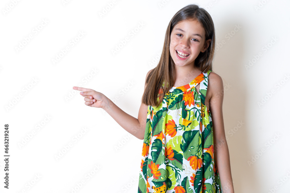 Beautiful kid girl wearing floral dress laughs happily points away on blank space demonstrates shopping discount offer, excited by good news or unexpected sale. Beautiful kid girl wearing floral dress laughs happily points away on blank space demonstrates shopping discount offer, excited by good news or unexpected sale.
