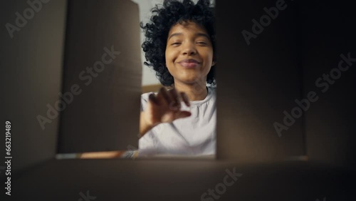 Young happy african american woman open cardboard box and looks at toothy smile with delight. Home lifestyle. Bottom view at portrait cute lady. Concept unpacking, product delivery and movement. photo