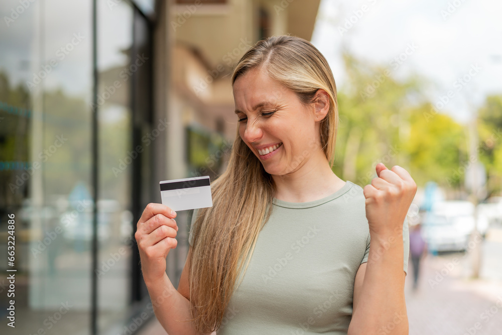 Young blonde woman holding a credit card at outdoors celebrating a victory
