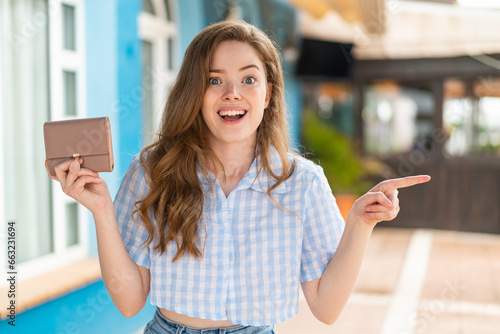 Young redhead woman holding a wallet at outdoors surprised and pointing finger to the side