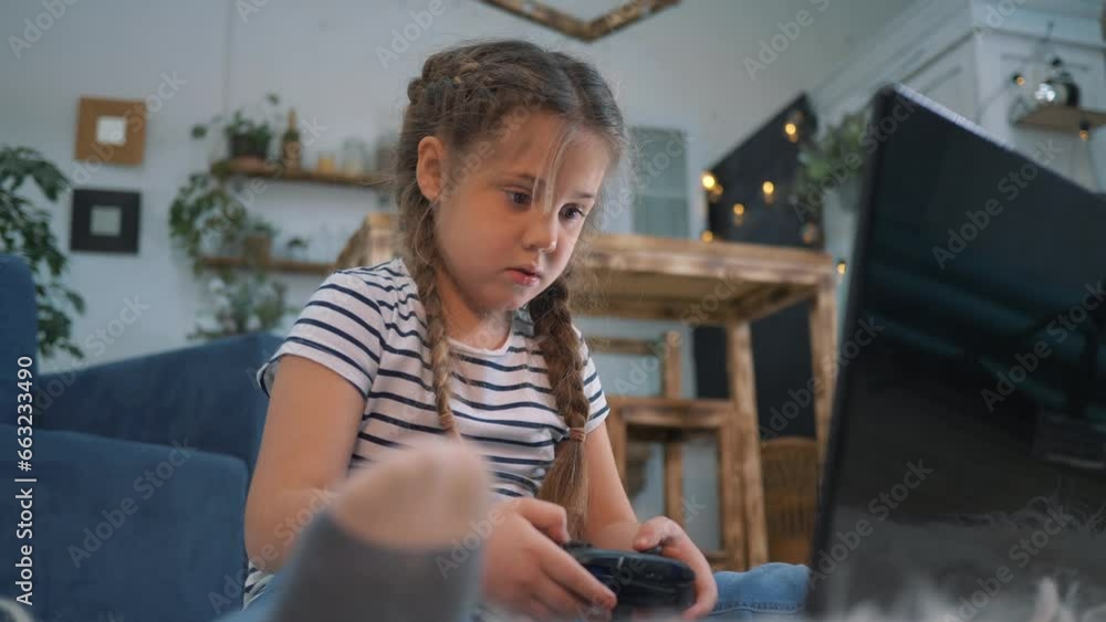 Happy toddler girl playing video games at home. Child controls game controller. girl gamer with help of control panel plays a computer game. Video game console for playing at home Happy toddler girl playing video games at home. Child controls game controller. girl gamer with help of control panel plays a computer game. Video game console for playing at home