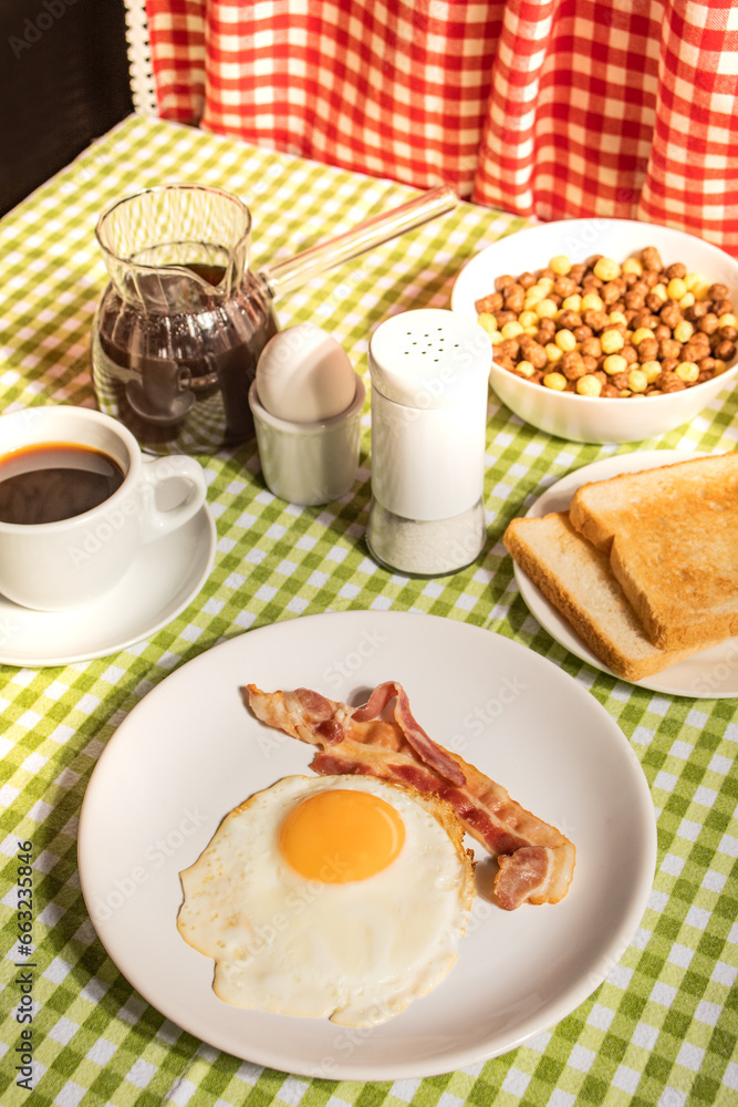 Fried egg with roasted bacon, black coffee, toasts on checkered tablecloth. Retro American breakfast. Hard shadow. Fried egg with roasted bacon, black coffee, toasts on checkered tablecloth. Retro American breakfast. Hard shadow.
