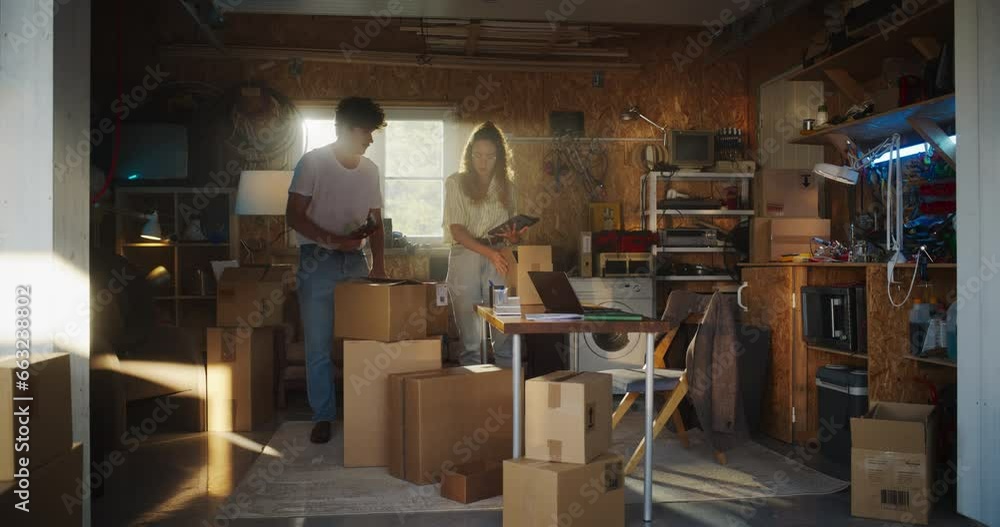Two Diverse Friends Working On Small Ecommerse Business In Garage. Female Hispanic Manager Using Tablet To Check Inventory, Caucasian Male Colleague Packing Products In Boxes For Online Customers. Two Diverse Friends Working On Small Ecommerse Business In Garage. Female Hispanic Manager Using Tablet To Check Inventory, Caucasian Male Colleague Packing Products In Boxes For Online Customers.