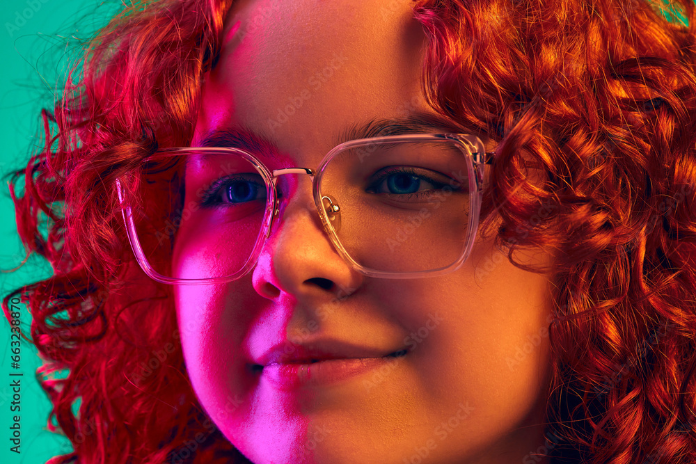 Close-up portrait of redhead teen girl with curly hair, in glasses, smiling against cyan background in neon light. Concept of human emotions, lifestyle, youth, facial expression. Ad Close-up portrait of redhead teen girl with curly hair, in glasses, smiling against cyan background in neon light. Concept of human emotions, lifestyle, youth, facial expression. Ad