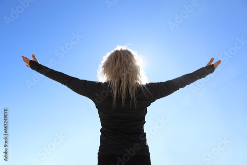 hands spread to sides. female Hiker arms outstretched on mountain top. woman on blue sky background. woman on peak. back, rear view of adult blonde lady. sporty clothing. backlit.