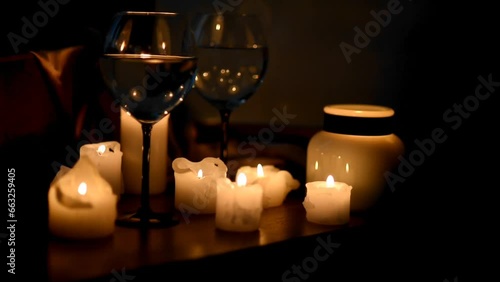Glasses of wine, burning candles in the dark. The flame is reflected by the glare in the glasses. Women's feet pass by. A woman picks up clothes, in the background. Romance, candlelit dinner