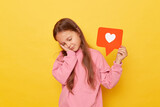 Sleepy tired little girl wearing pink sweatshirt holding blogger heart like icon keeps eyes closed leaning on her hands having nap posing isolated over yellow background.
