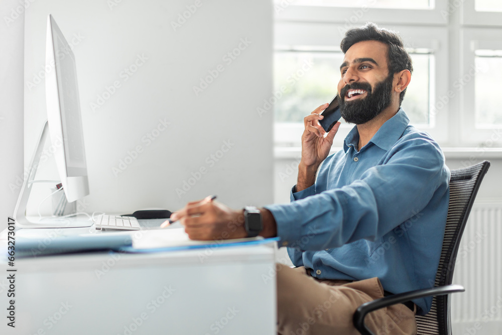 Positive thoughtful indian businessman talking on cellphone and taking notes while working on computer in office - obrazy, fototapety, plakaty 