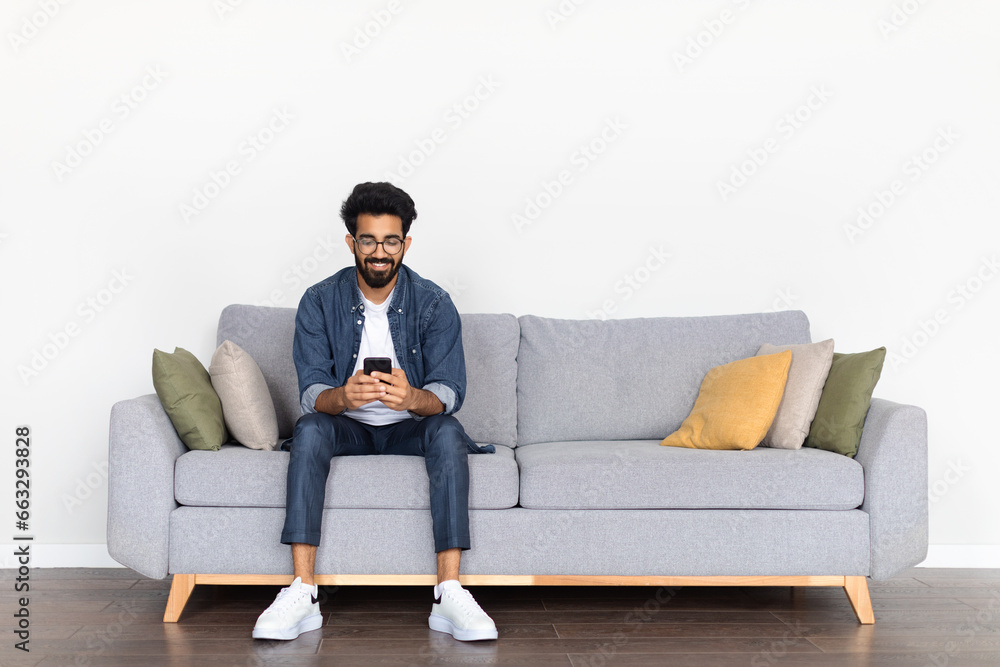 Casual Indian Male Enjoying Time on Couch with Phone - obrazy, fototapety, plakaty 