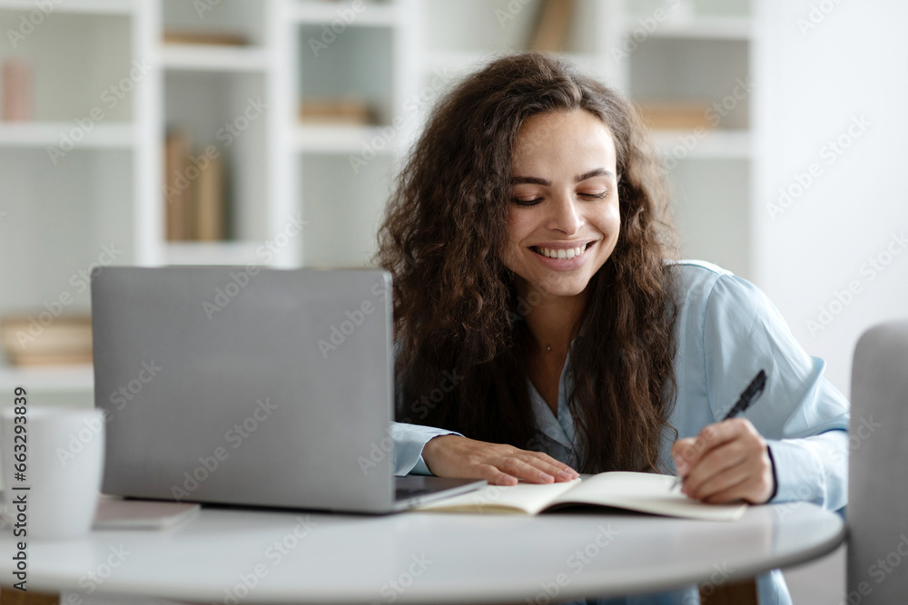 Distance learning. Happy lady studying with laptop and taking notes, writing in notepad and using computer sitting at table in living room at home Distance learning. Happy lady studying with laptop and taking notes, writing in notepad and using computer sitting at table in living room at home