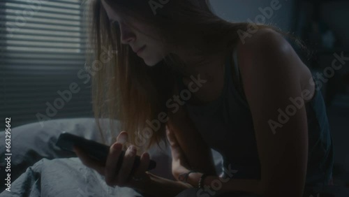 Tilt up shot of nervous girl sitting in dark bedroom, doomscrolling on phone and feeling anxiety during night