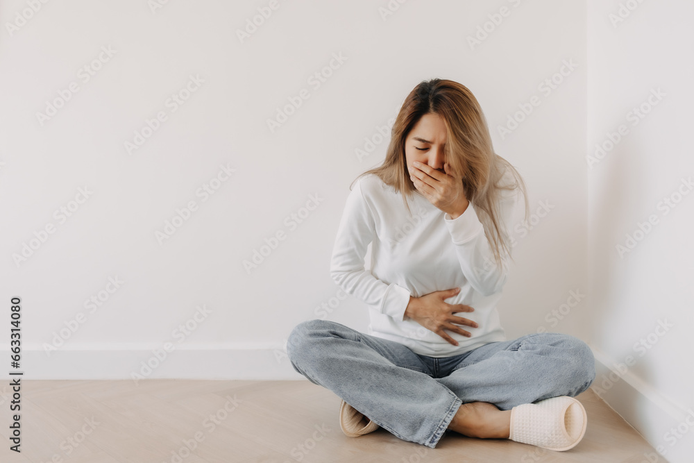 Asian Thai woman feeling sick and queasy, nausea and vomiting, grabbing stomachache cramp and covering mouth, wear white sweater, sitting on floor in white apartment room in winter.