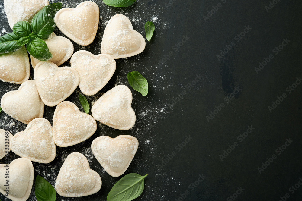 Italian ravioli pasta in heart shape. Tasty raw ravioli with flour and basil on dark background. Food cooking ingredients background. Valentines or Mothers Day lunch ideas. Top view with copy space. Italian ravioli pasta in heart shape. Tasty raw ravioli with flour and basil on dark background. Food cooking ingredients background. Valentines or Mothers Day lunch ideas. Top view with copy space.