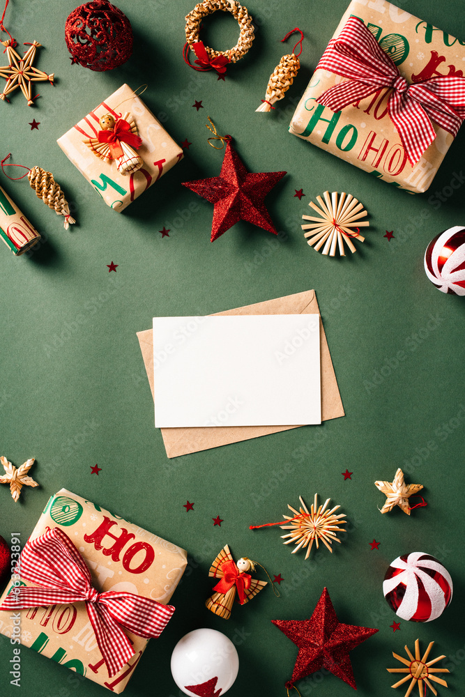 Fototapeta premium Christmas letter to Santa concept. Top view blank paper card with envelope on green background decorated red and white balls, handicraft gift boxes, rattan decorations. Vintage, retro style.