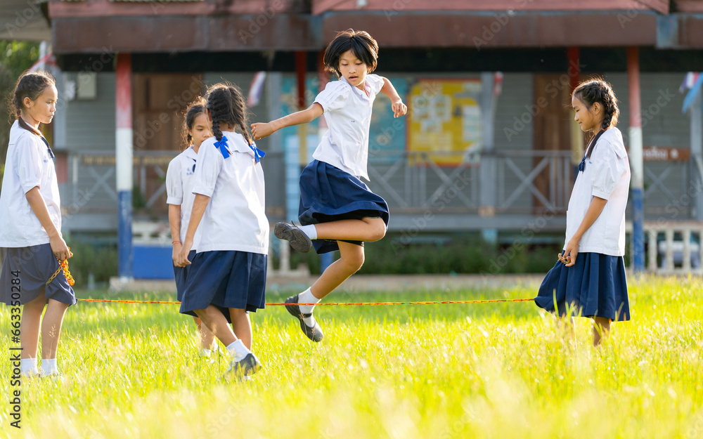 Fototapeta premium Close up group of Asian girls wear student uniform enjoy to play with jump across rubber string in grass field area of school.