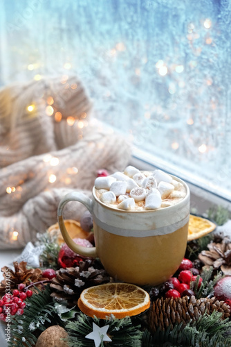 New year and Christmas holidays background. coffee mug with marshmallows, festive decor, sweater, fir branches close up on window sill. frozen window. festive winter season.