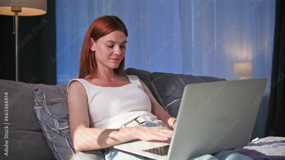 remote work, charming young woman working at computer while sitting on sofa at home, smiling and looking at camera remote work, charming young woman working at computer while sitting on sofa at home, smiling and looking at camera