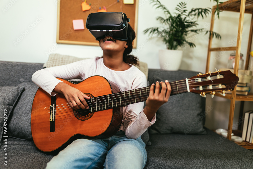 Obraz premium African american teenage girl sitting on couch in her room and learning to play guitar using VR glasses to immerse herself in metaverse