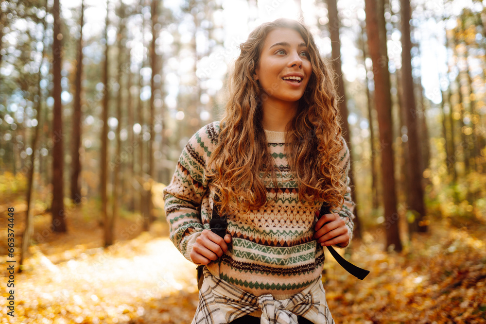 Beautiful young tourist with a backpack is having fun in the autumn forest, throwing fallen leaves. Woman walks along a colorful path in the forest. Freedom, travel. - obrazy, fototapety, plakaty 