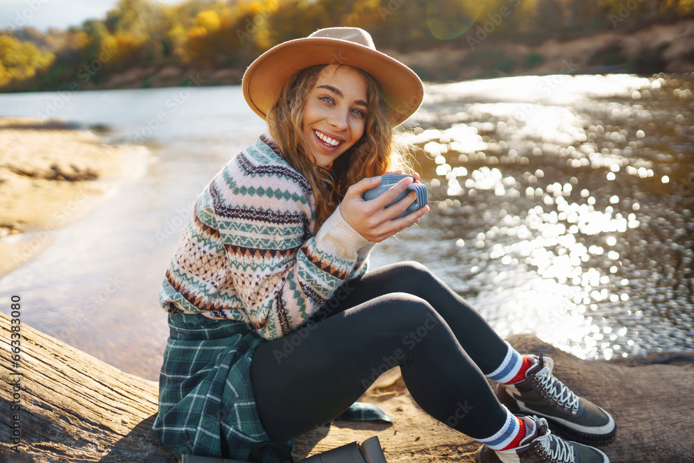 Young woman traveler in a hat enjoying autumn nature on the river bank, drinking a hot drink from a thermos. Beautiful woman drinking tea, sitting by the lake outdoors.