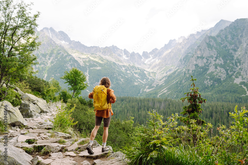 Smiling traveler with a bright backpack on a hiking mountain trail. A beautiful woman on a cliff enjoying the mountain scenery. Active lifestyle.