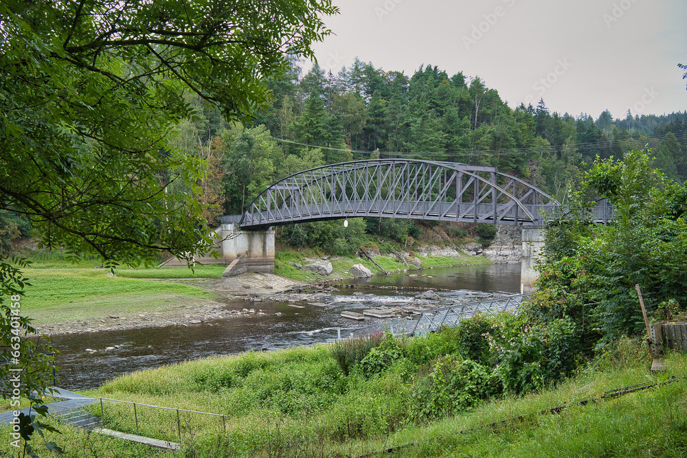 Fototapeta premium Seitenansicht der stählernen Fachwerkbogenbrücke über die Saale bei Harra mit Blick auf das bewaldete Ufer und das steinige Flussbett, Thüringen, Deutschland