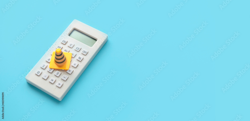 Concept of under construction or fixing for financial, loan, or budget. Safety cone or traffic cone over a calculator on blue background with copy space. Concept of under construction or fixing for financial, loan, or budget. Safety cone or traffic cone over a calculator on blue background with copy space.