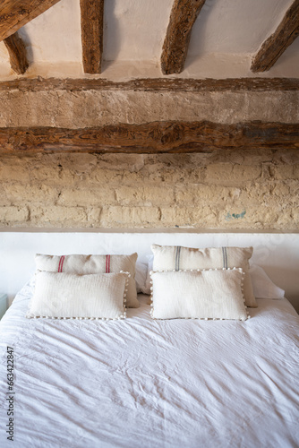 interior of rustic rural room, bed with white sheets and cushions, front view of headboard with white shelf and rustic stones of European rural house.