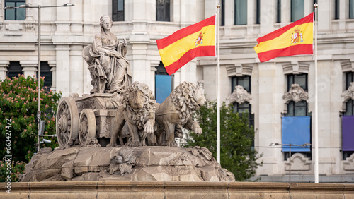 Statue in the Plaza de Cibeles with the representation of the goddess of the same name and two lions with Spanish flags waving in the wind behind photo