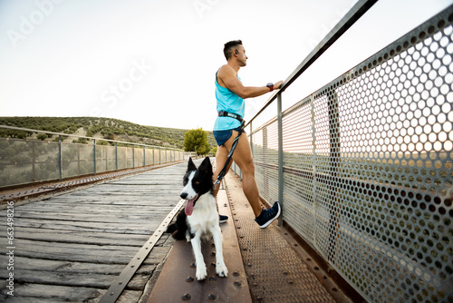 Foto An adult runner with an amputated arm stretches his calf leg on an outdoor bridge fence at sunset with his dog
