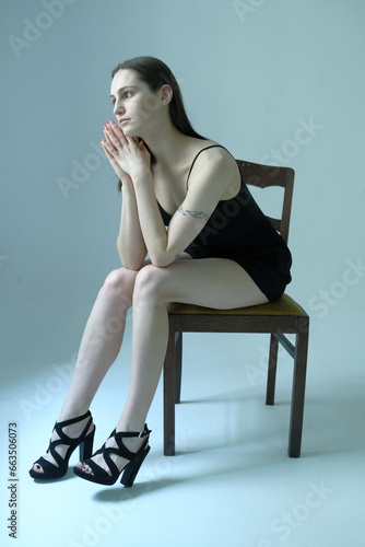 Portrait of a brunette girl without make up on a grey backgroung in a black summer clothes. Woman is sitting on a chair