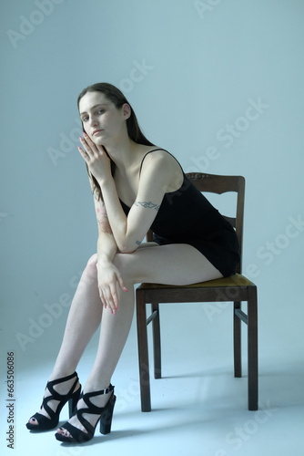 Portrait of a brunette girl without make up on a grey backgroung in a black summer clothes. Woman is sitting on a chair